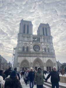 Photo of the Notre Dame from a distance, with many people standing at the base. Cloudy sky in the background.