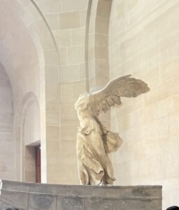 Statue of the Winged Victory of Samothrace, a headless and armless marble figure with wings, displayed on a stone base in a museum with arched walls.