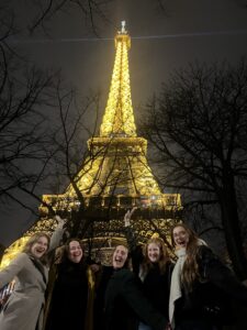 Five smiling people pose excitedly in front of the glowing Eiffel Tower at night, framed by bare tree branches.
