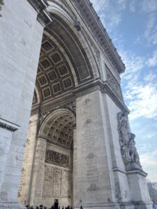 Photo of the Arc de Triomphe in Paris, showing its large stone arch, detailed carvings, and engraved names. People stand at the base, and the sky is partly cloudy.