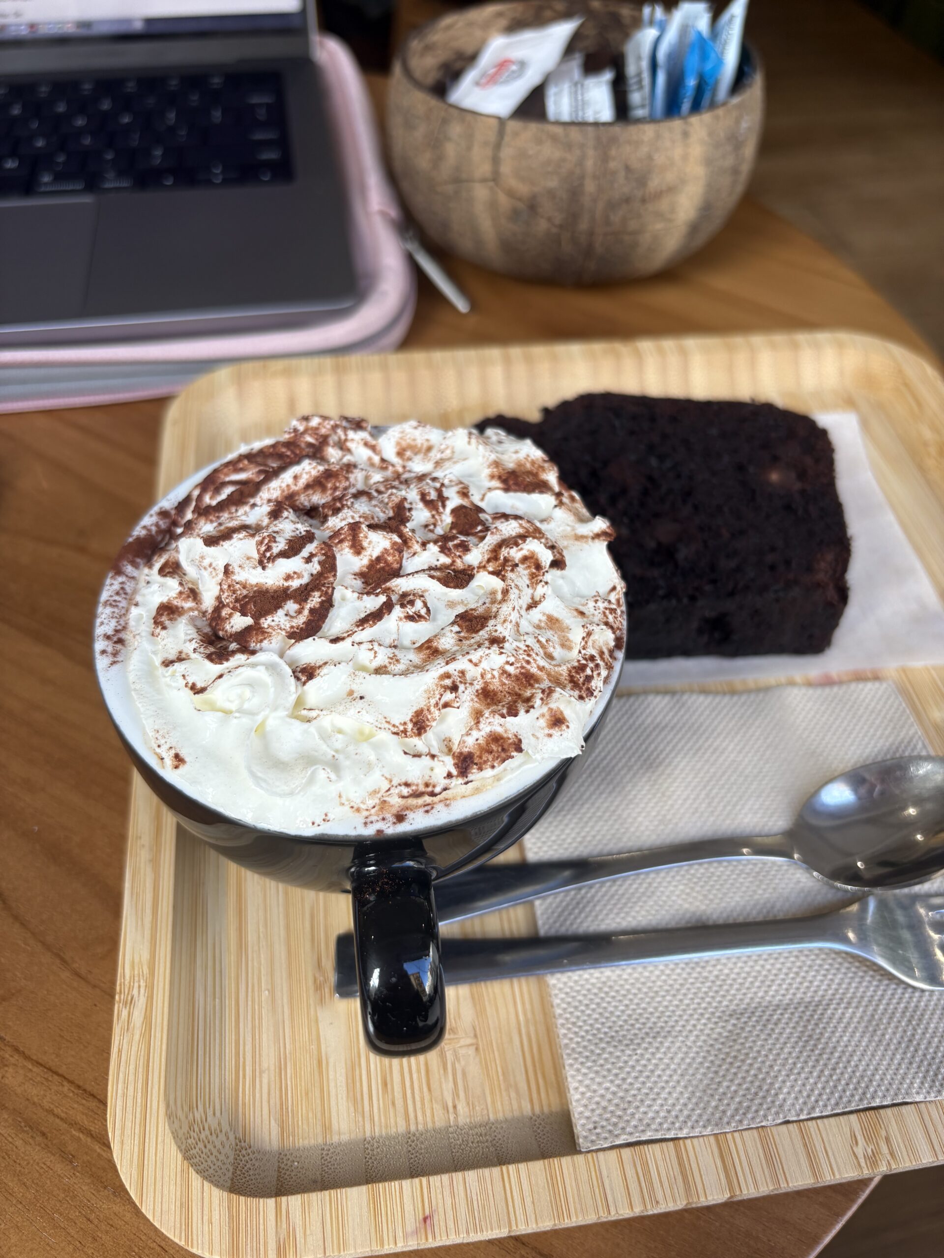 A photo of a mocha in a black mug, with whipped cream and cocoa powder on too, a silver spoon and fork, and chocolate chip banana bread on a beige wood tray, with my silver computer and pink computer case in the background, as well as a brown bowl with sugar packets inside, all on a brown wood table