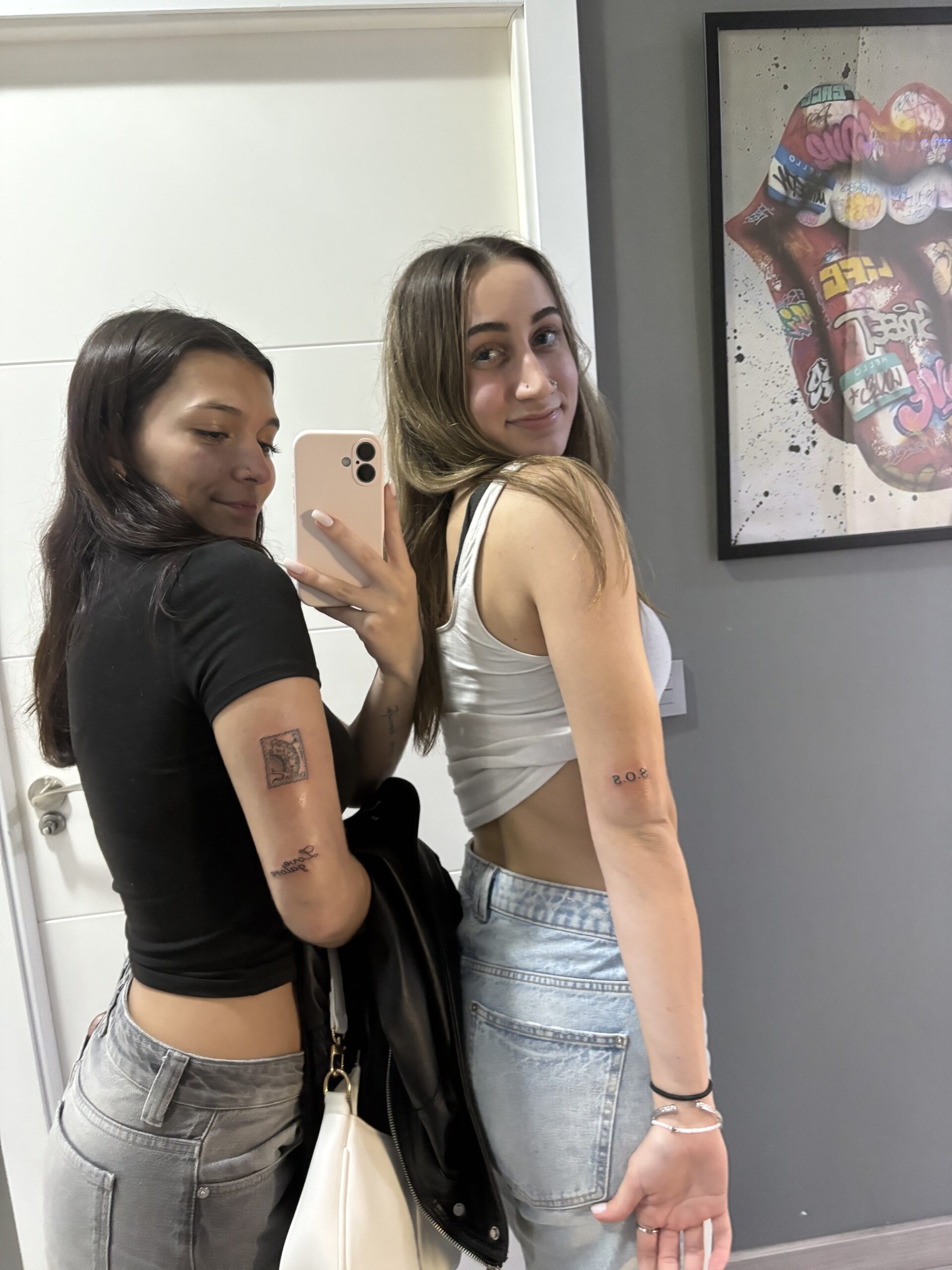 A photo of me and my friend Erica right after getting matching tattoos on the back of our arms above the elbow, mine saying SOS and hers saying Love Language with a Spanish stamp above it, I'm wearing a white top and blue jeans and she's wearing a black top and gray jeans, with a white door in the background as well as a picture frame of a painted colorful mouth with the tongue sticking out
