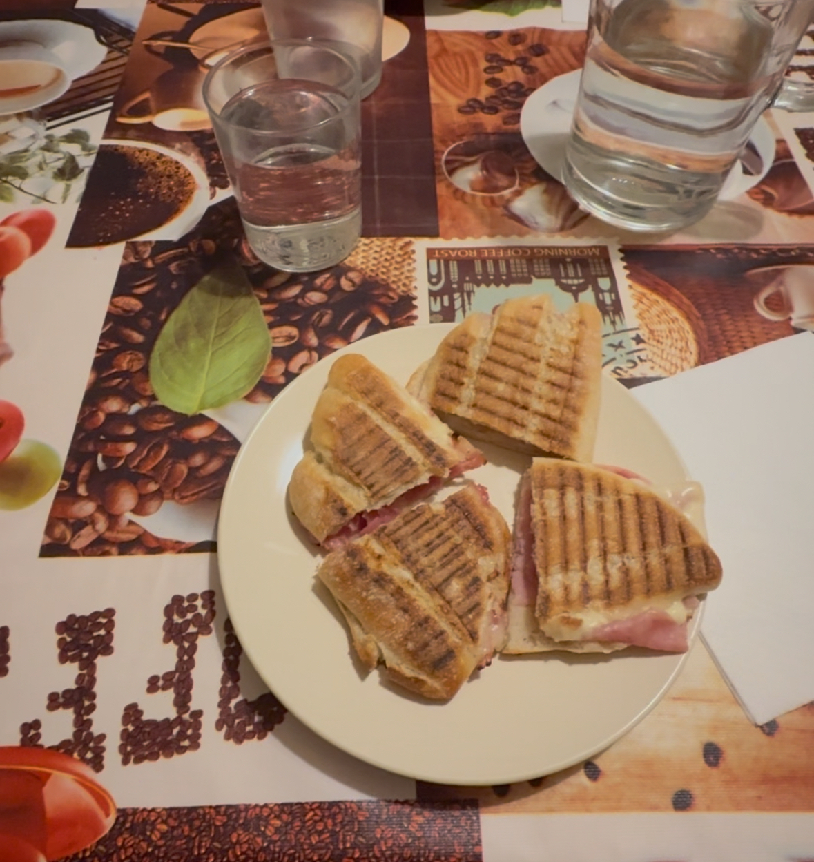 A photo of one of my dinners on a table with a coffee themed table cloth, with two glasses of water and a beige ceramic plate with a grilled panini