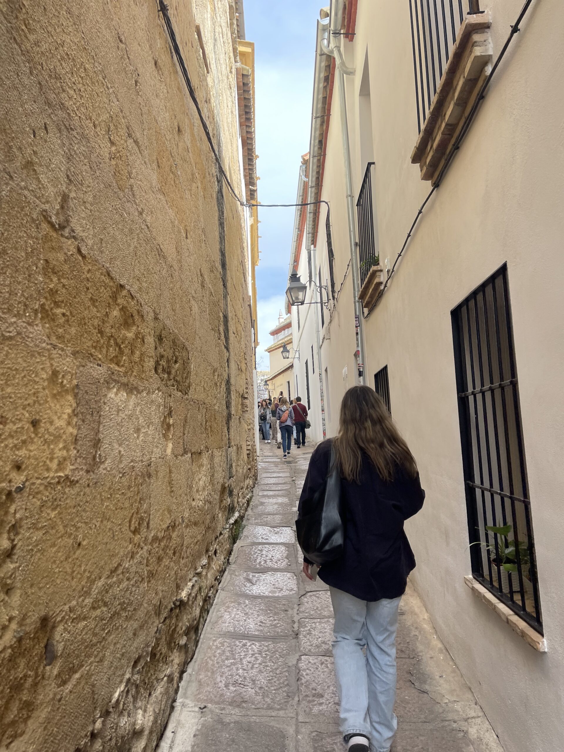 Me walking through an alley in Córdoba wearing blue jeans, a dark navy jacket and a black leather bag, there are two narrow walls on either side of me with a beige colored stone, with black metal grated windows, with a sliver of blue sky peeking out at the top