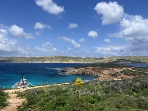 View of a bright blue sea and rocky coastline, with a small boat docked and wildflowers in the foreground under a partly cloudy sky.