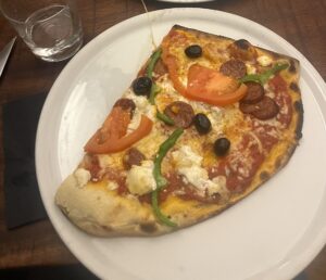 Large pizza slice on a wooden table, topped with tomato, green pepper, olives, sausage, and cheese. Glasses and utensils are also on the table