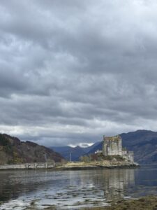 A castle on a peninsula with mountains behind it and a the sea in front.  Lots of clouds in the sky.