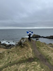 Girl in navy blue jacket waves Scottish flag while standing .on the edge of a cliff with beautiful views of the ocean.