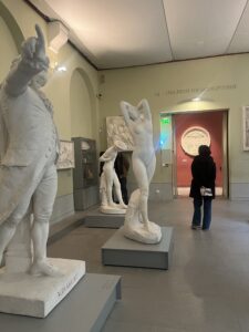 A museum gallery with several white marble sculptures on display. In the foreground, a statue of a man in historical attire points upward with his right hand, labeled “MIRABEAU.” Behind it, a nude female statue stands with her arms raised behind her head, and another partially visible statue appears further back. Two visitors stand near the entrance at the back, one facing the statues and the other facing a red-walled room with a round artwork. 