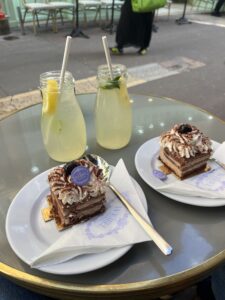 Two pieces of chocolate cake sit on plates. Whipped cream, brown powder, a cherry, and a circular purple paper labeled Weibel sit on top of the cakes. Behind them are two jars of lemonade with mint and lemons slices inside the glasses. They all sit on a circular table with gold trim.