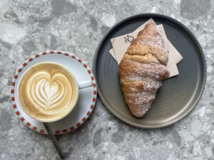 A croissant with powdered sugar dusted on top sits on a grey plate. To the left is a latte with a heart design in the foam sitting on a smaller white plate with red and green trim. The two plates sit on a grey marble table. 